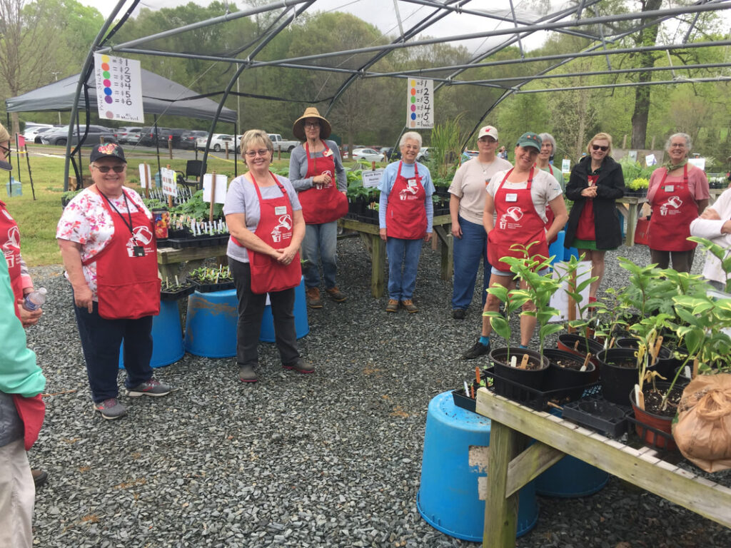 8 people wearing red aprons standing among plants that are on tables