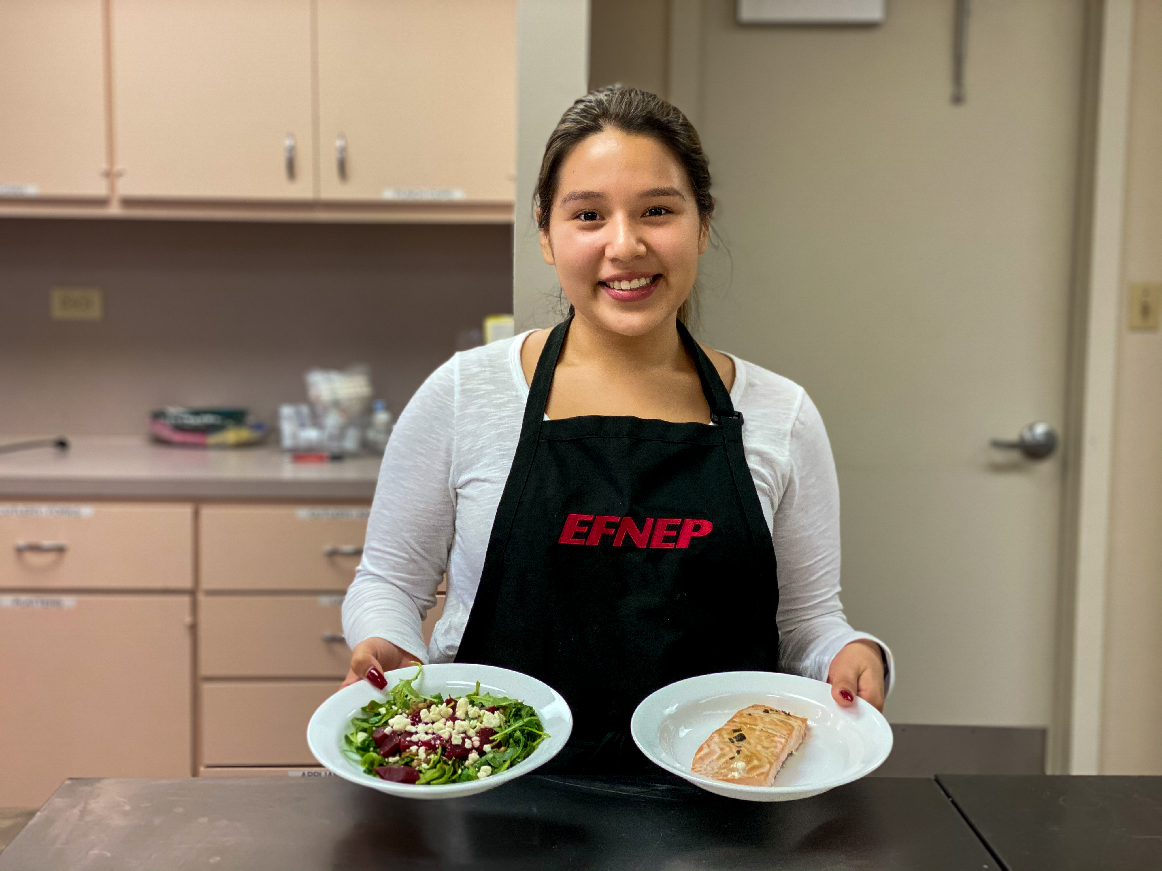 Woman in kitchen wearing black apron and holding two plates. 